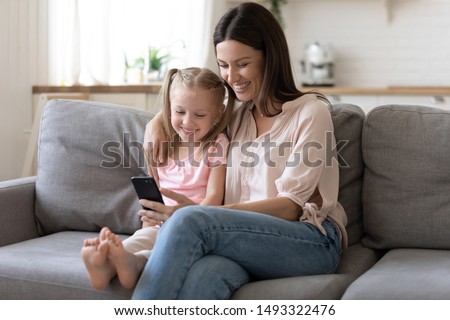 Mother embraces little preschool daughter sitting on sofa mom holding in hands smartphone family smiling looking at screen using new application, e-commerce buying online have fun in internet concept