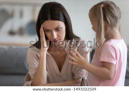 Annoyed mom and restless little daughter at home, mother sitting on couch touch head or temples feels headache due to noisy kid standing near her, tired parent of difficult hyperactive child concept Royalty-Free Stock Photo #1493322509