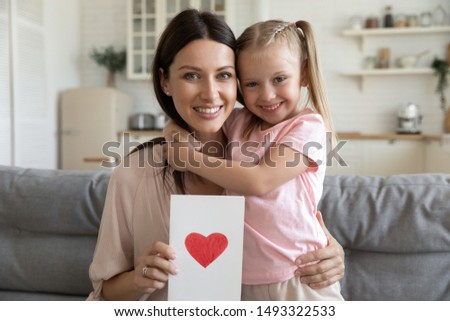 Happy beautiful woman holding postcard with drawn red heart embraces little daughter people sitting on couch at home looking at camera, concept of life events celebration, family holidays, mothers day Royalty-Free Stock Photo #1493322533