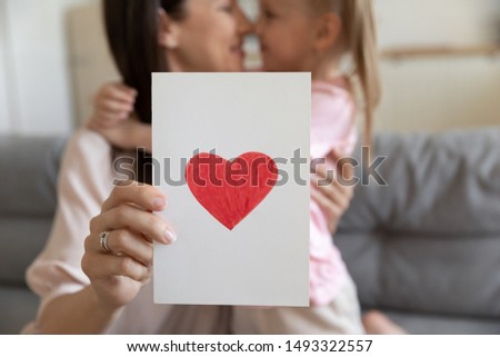 Close up focus on white paper handmade with love postcard with red drawn heart holding mommy embraces little daughter, mom express gratitude kiss lovely attentive kid, mothers day celebration concept