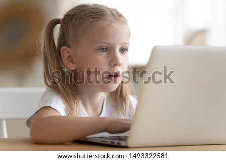 Close up of little girl sit at table looking at pc screen spends time watching cartoons or play game on laptop, younger generation addicted with virtual amusements, computer overuse, bad habit concept