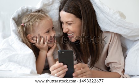 Mother daughter using smartphone laughing lying in bed under blanket close up image, enjoy free time in internet online chat, interesting education program users amusement kids and modern tech concept
