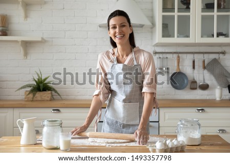 Woman wearing apron smiling looking at camera standing in kitchen preparing for family dinner holding rolling pin flattening dough eggs flour milk on wooden table, housewife chores or hobby concept Royalty-Free Stock Photo #1493357798