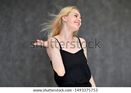 Portrait of a cute blonde student girl, a young woman in a black dress with beautiful curly hair on a gray background in different poses. Shows hands to the sides. Beauty, brightness, emotions.