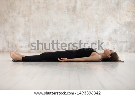 Young woman wearing black sportswear practicing yoga, doing Corpse, Savasana exercise, relaxing, lying in Dead Body pose on mat, sporty girl working out at home or in yoga studio with grey walls Royalty-Free Stock Photo #1493396342