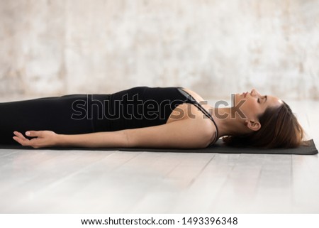 Young woman wearing black sportswear practicing yoga, doing Corpse, Savasana exercise, relaxing, lying in Dead Body pose on mat, girl with closed eyes working out at home or in yoga studio close up Royalty-Free Stock Photo #1493396348