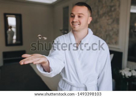 Stylish groom in a terry dressing gown throws rings and catches. Wedding portrait of a smiling man. Concept and photography.