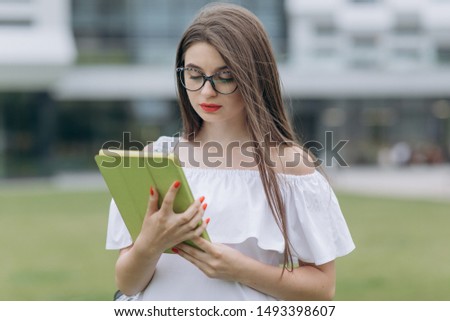 Portrait from below on the young pretty businesswoman using tablet device while standing outdoors big urban building