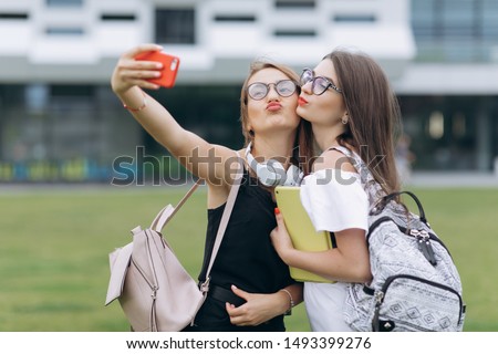 Outdoor portrait of pretty happy girls making video call to her best friend relaxing in the courtyard. Female bloggers filming a selfie video in modern city background.