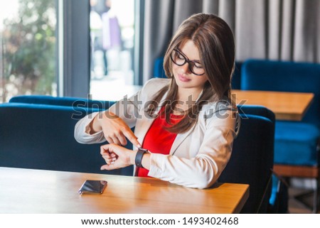 Portrait of beautiful stylish brunette young woman in glasses sitting checking notification or message and touching her smart watch. indoor studio shot, cafe, office background.