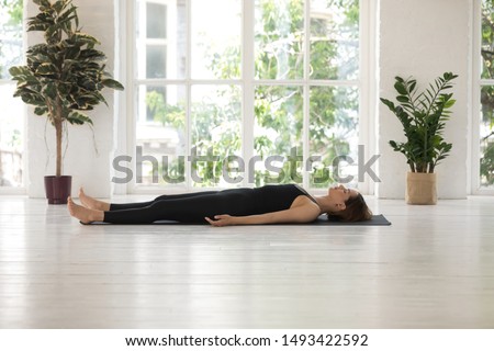 Young woman wearing black sportswear doing Savasana exercise, relaxing in Dead Body, Corpse pose, practicing yoga, beautiful sporty girl working out at home or in yoga studio with window and plants Royalty-Free Stock Photo #1493422592
