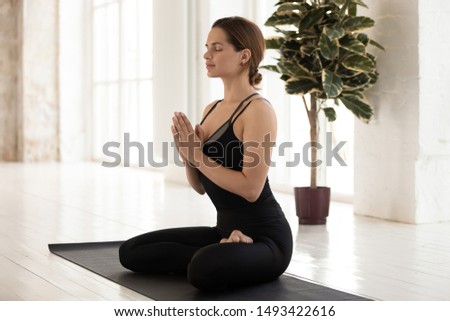 Young woman with closed eyes relaxing, sitting in Lotus pose on mat, doing Padmasana exercise, practicing yoga, attractive sporty girl wearing black sportswear working out at home or in yoga studio Royalty-Free Stock Photo #1493422616