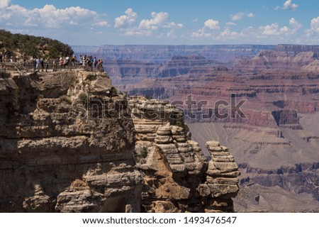 A group tourists in colorful clothing looking tiny in comparison to the huge rock formation they are standing on as they look out over a giant ravine that makes up part of Grand Canyon National Park.