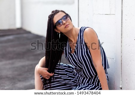 Portrait of young brunette woman with black afro american dreadlocks hairstyle in striped dress and sunglasses, sitting, posing, holding her hairs and looking at camera. fashion model shot.
