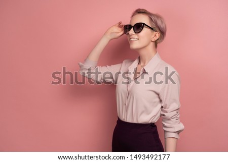 A girl with short pink hair in a blouse, skirt and glasses stands in a half-turn on a pink background and looks to the top.