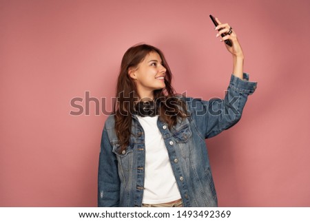 A dark-haired girl in a jeans stands on a pink background with headphones on her neck and takes a selfie.