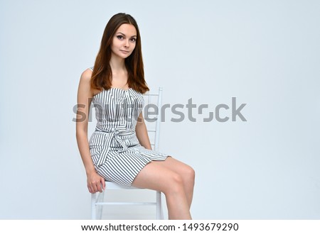 Portrait of a cute brunette girl with beautiful curly hair in a gray dress sitting on a chair on a white background. Smiling, talking with emotions.