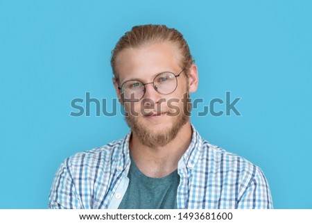 Young bearded handsome man blond hair hipster face wearing optical glasses looking at camera, millennial handsome casual guy isolated on blue studio blank background, close up view headshot portrait