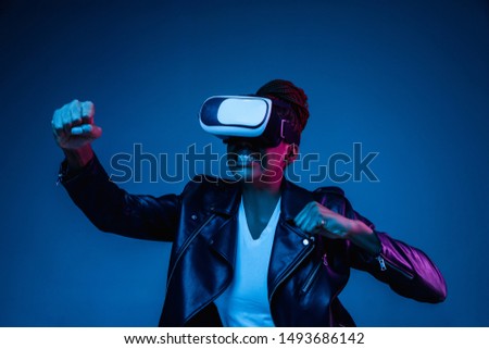 Portrait of young african-american woman's playing in VR-glasses in neon light on blue background. Concept of human emotions, facial expression, modern gadgets and technologies. Look fighting, angry.