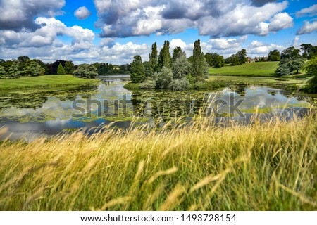 River Glyme in Oxfordshire UK. Lake dammed in river with small tree covered island. Natural environment. Summer landscape with sunshine and blue skies. Royalty-Free Stock Photo #1493728154