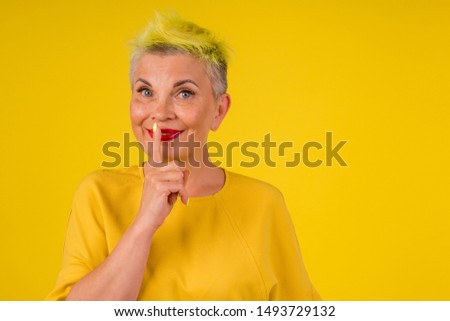kind positive mature woman with colored stylish yellow dyed hair in warm dress and make up red lipstick on lips shh gesture background studio copy spase.anti-aging face gymnastic.