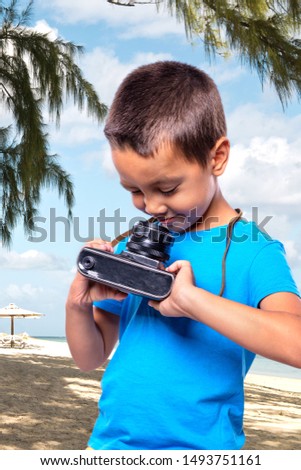 A little boy wearing blue shirt is looking at the camera curiously.  He is standing on the beach and about to take a picture.