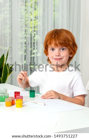 Little red-haired boy sits at the table in the room and  paint with colors.  Hi is looking at camera and smiling