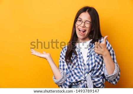 Picture of a happy pleased smiling young cute teenage girl in glasses posing isolated over yellow wall background showing copyspace make thumbs up.
