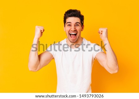 Positive brunette man rejoices in victory. Portrait of guy in white T-shirt on orange background