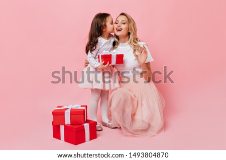 Curly daughter kisses fair-haired mother, receiving birthday present. Portrait of lovely adult lady and little girl in light dresses on pink background