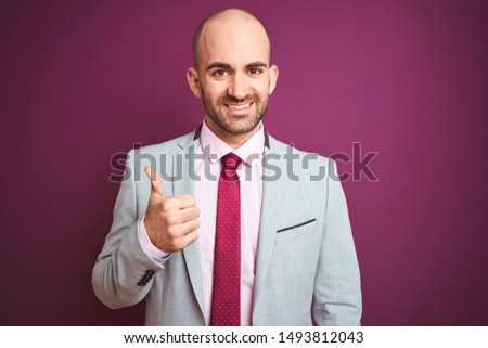 Young business man wearing suit and tie over purple isolated background doing happy thumbs up gesture with hand. Approving expression looking at the camera with showing success.