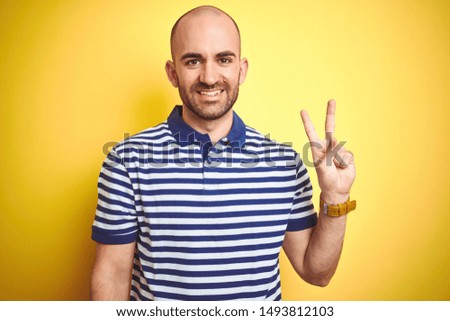 Young bald man with beard wearing casual striped blue t-shirt over yellow isolated background showing and pointing up with fingers number two while smiling confident and happy.