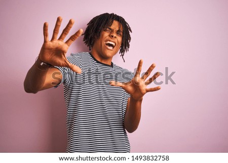 Afro man with dreadlocks wearing navy striped t-shirt standing over isolated pink background afraid and terrified with fear expression stop gesture with hands, shouting in shock. Panic concept.