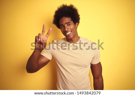 American man with afro hair wearing striped t-shirt standing over isolated yellow background smiling looking to the camera showing fingers doing victory sign. Number two.