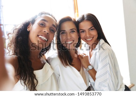 Image of smiling positive amazing pretty young women colleagues at workplace indoors in office take a selfie by camera.