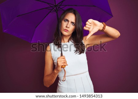 Young beautiful woman holding umbrella standing over purple isolated background with angry face, negative sign showing dislike with thumbs down, rejection concept