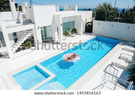 fashion outdoor photo of beautiful girl with dark hair in beach clothes posing at luxurious white villa with swimming pool