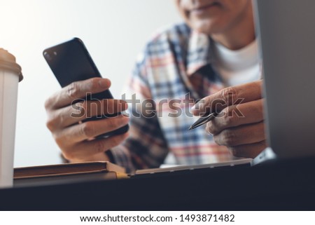 Casual business man reading E-book on mobile apps via smart phone, connecting internet while working on laptop computer in office. Freelance networking on smartphone. E-learning, online business 