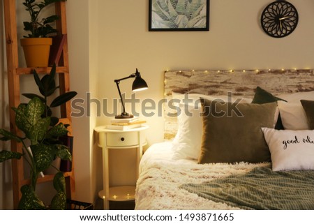 Big comfortable bed and table with lamp and plants in stylish interior of room