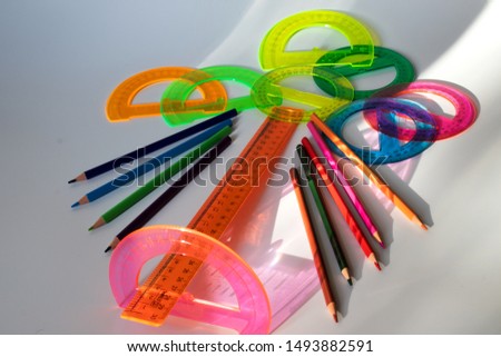 Pink protractor with a measuring ruler lies on a white background. In the background are colored pencils and round, colored protractors. On the table are glare from the sun.
