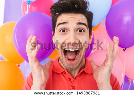 Image closeup of joyful brunette man rejoicing with glitter stars on his face and multicolored air balloons isolated over purple background