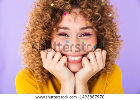Image closeup of attractive curly girl having fun and smiling with multicolored glitter stars on her face isolated over violet background