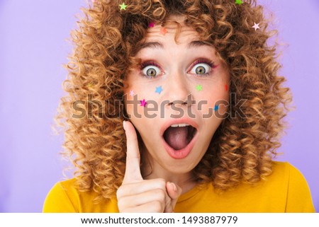 Image of pretty curly girl having idea and pointing finger upward with multicolored glitter stars on her face isolated over violet background