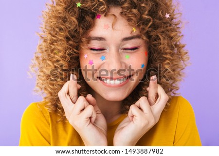 Image closeup of optimistic pretty curly girl keeping fingers crossed with multicolored glitter stars on her face isolated over violet background