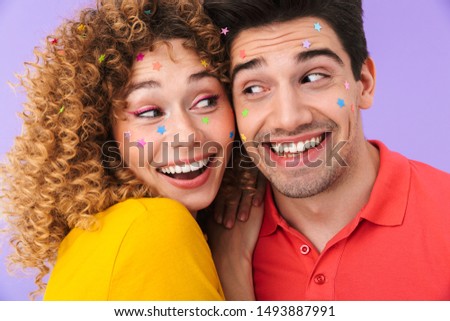 Image of young attractive couple smiling and hugging while having party with glitter stars on faces isolated over violet background