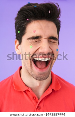 Image closeup of caucasian delighted man having fun and screaming with multicolored glitter stars on his face isolated over violet background