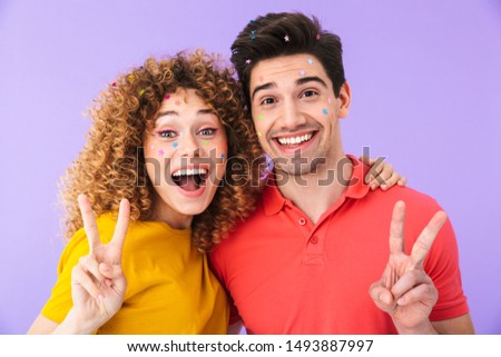 Image of beautiful couple smiling and showing peace fingers while having party with glitter stars on faces isolated over violet background