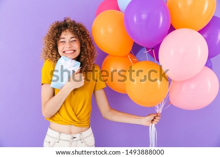 Image of happy young woman celebrating birthday with multicolored air balloons and present box isolated over violet background
