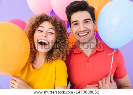 Image of happy couple man and woman smiling at camera while standing with multicolored air balloons isolated over violet background