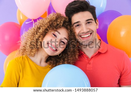 Image of beautiful couple man and woman smiling at camera while standing with multicolored air balloons isolated over violet background
